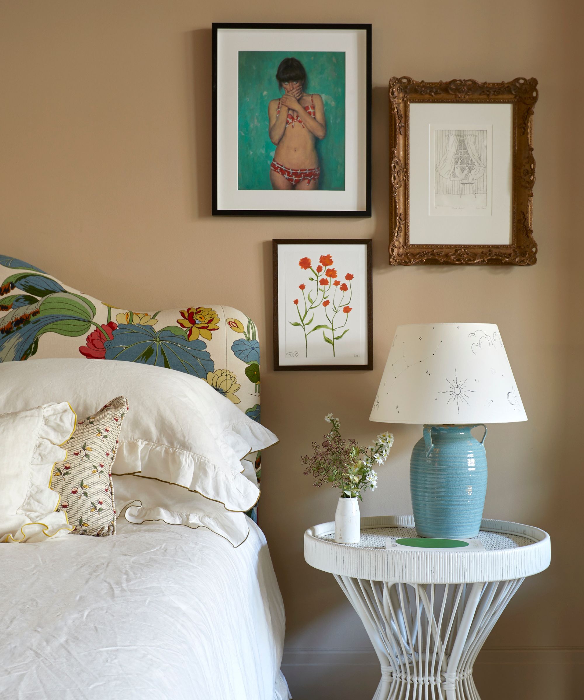 A close-up of an eclectic bedroom featuring a bed with a vibrant floral and lily pad patterned headboard. Next to the bed is a white wicker side table holding a light blue ceramic lamp with a hand-drawn celestial shade. The beige wall above is decorated with a small gallery of framed art, including a portrait of a woman in a red bikini and a botanical illustration of orange flowers.
