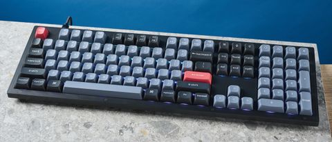 A photograph of the Keychron V6 in black, with gray , black and orange keycaps. The keyboard is positioned on a stone slate, with a blue wall in the background.