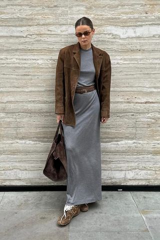 A woman standing against a marble wall outside wearing brown sunglasses, a brown suede blazer, a gray T-shirt dress, layered brown belts, suede sneakers, and an oversize suede tote bag.