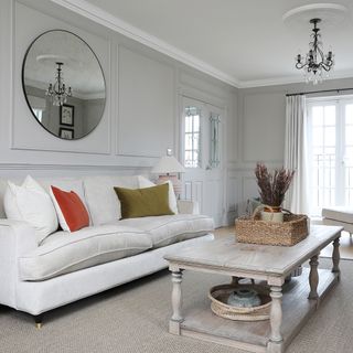 a neutral look living room with a cream sofa and velvet cushions, large wooden coffee table and wall panelling with an oversized round foxed mirror