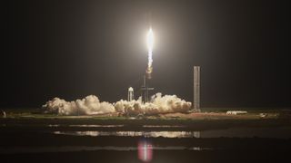 a white rocket lifts off at night above a plume of fire and smoke