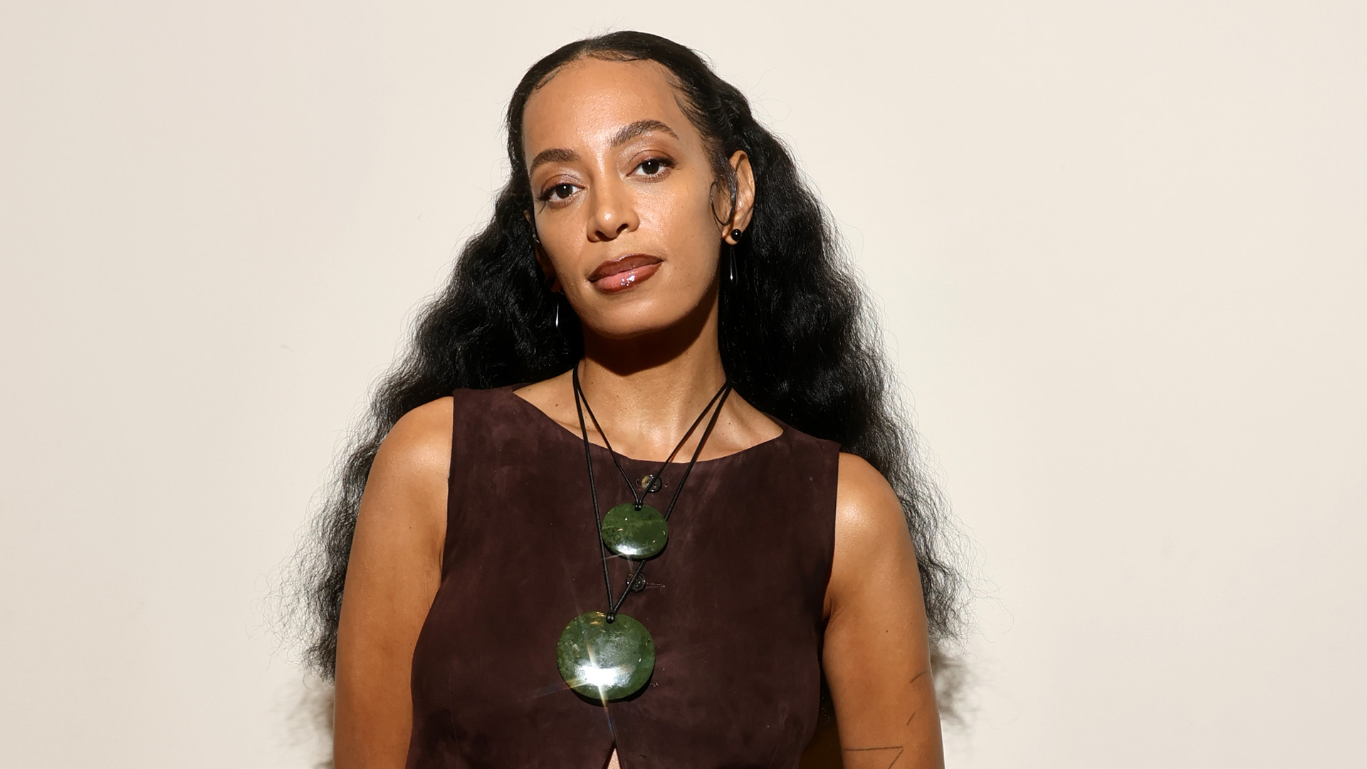 Solange Knowles (a woman with long, wavy hair) wearing a brown top and green necklaces looking at the camera.