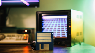 A floppy disk, visible against a background showing an old display and keyboard.