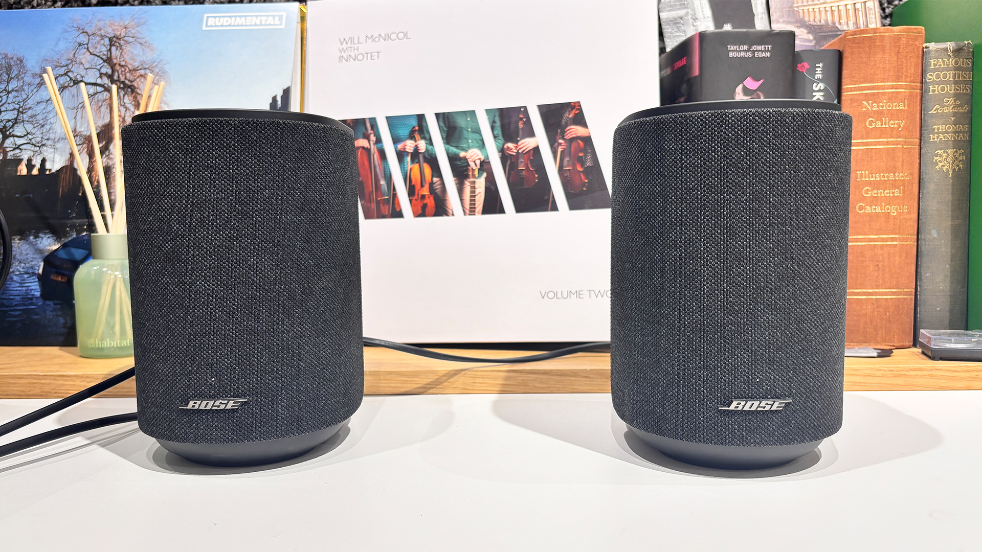 Two Bose Lifestyle Ultra Speaker wireless speakers on white surface in front of busy bookshelf