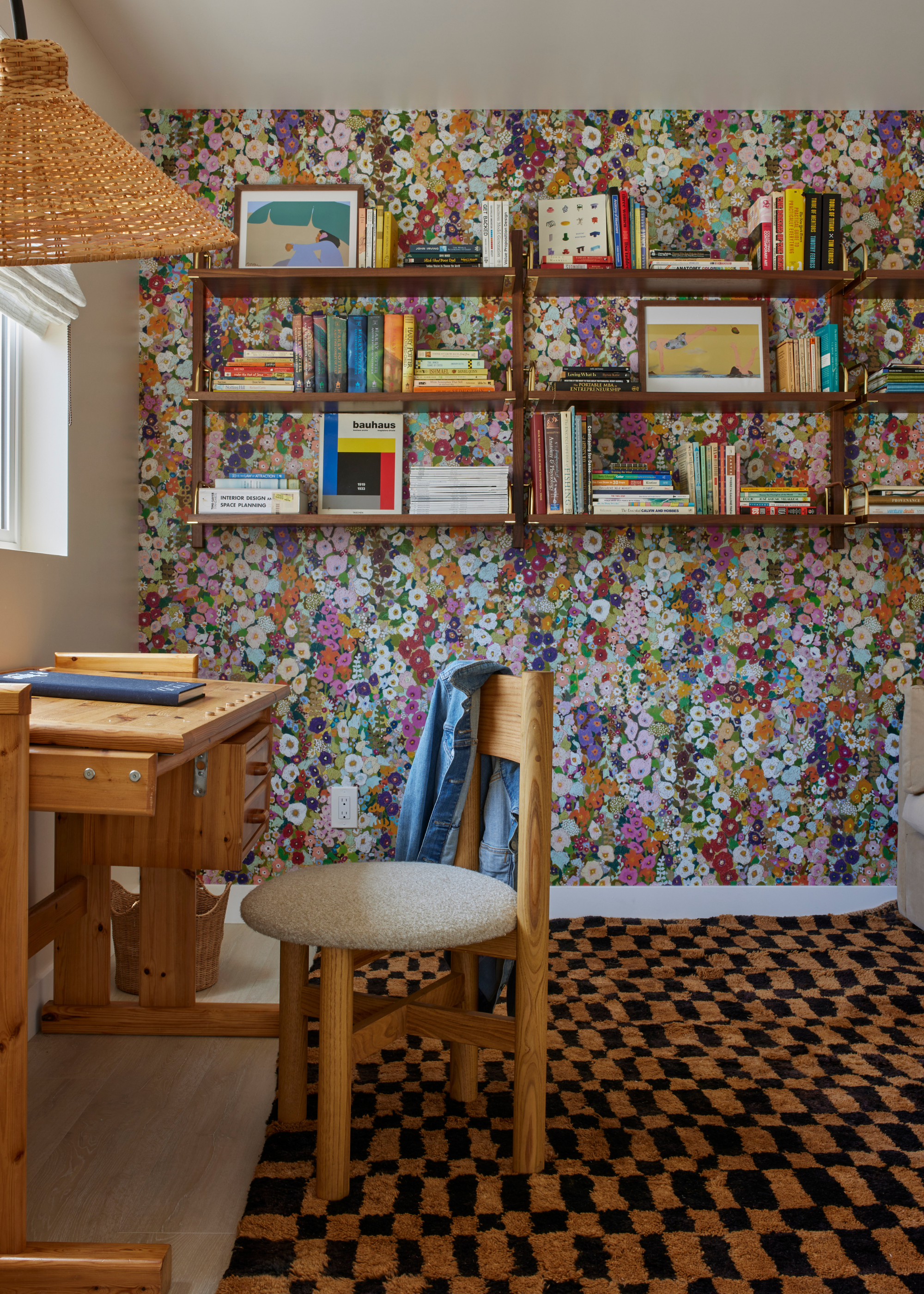 A study with a checkerboard carpet, a wooden desk and chair, floating shelves of books, a woven lamp shade, and floral wallpaper