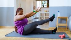 A woman sits on a Pilates mat at home with her arms lifted in front of her face. Her left leg is straight on the floor and her right leg is lifted at a 45° angle. Behind her we see a wooden stool and water bottle and a kitchen counter.