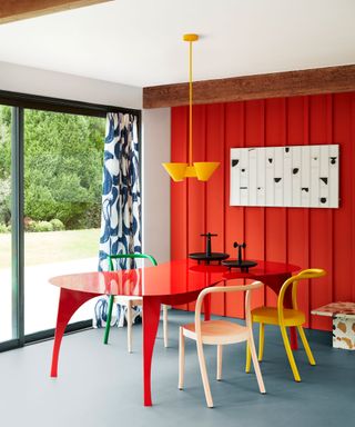 Modern dining room with bright red, painted paneled wall, curvaceous glossy red dining table, bright yellow, green and white dining chairs, low hanging yellow cone pendant, blue flooring, black and white artwork, blue and white patterned curtains, table positioned beside large french doors