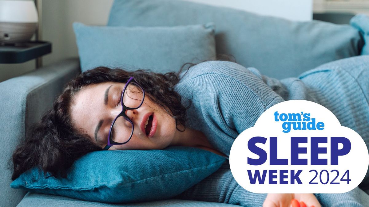 A woman with dark curly hair and glasses sleeps on her side on a blue sofa with her mouth open