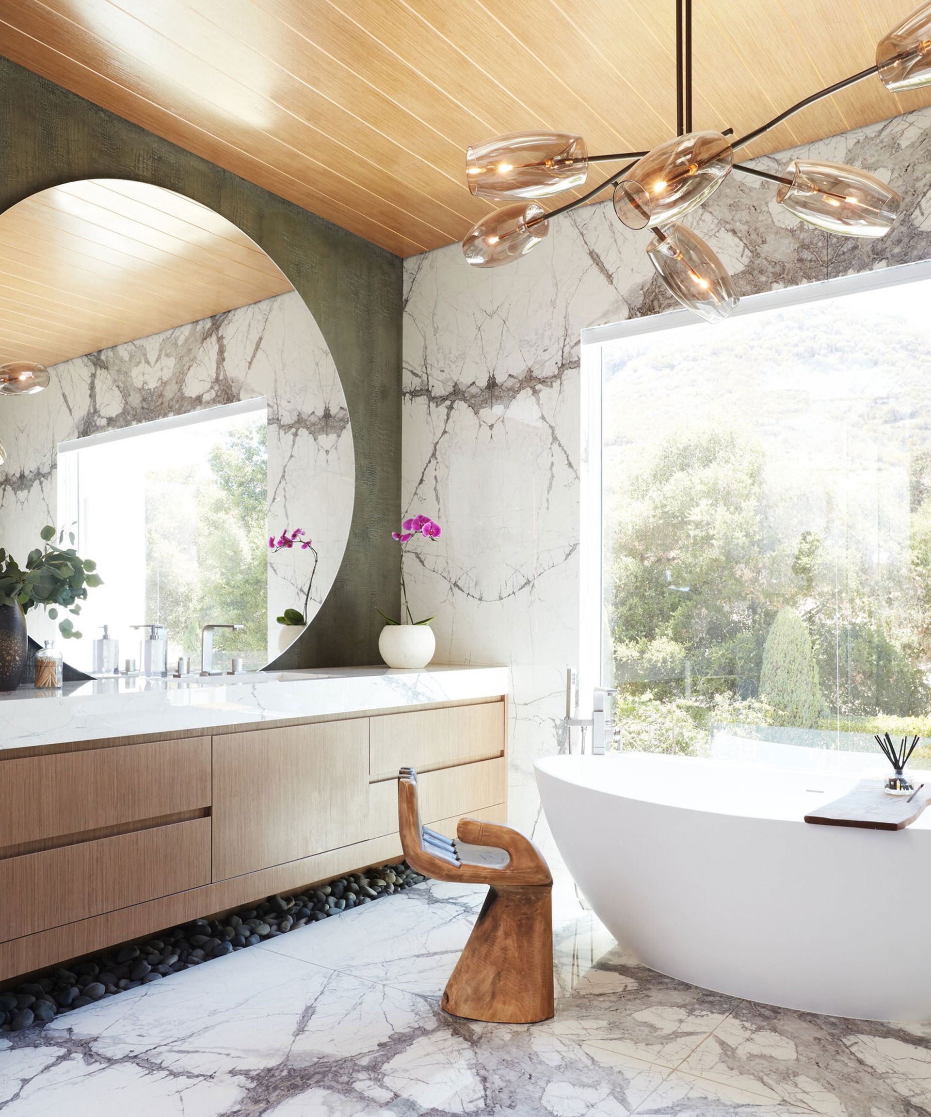 Large, modern bathroom with wooden paneled ceiling, modern, metal and glass chandelier, marble tiles on one wall, large white bath, wall to wall vanity unit with wooden draws, marble countertop, large round mirror above sinks, marble flooring, green textured wall, wooden stool in shape of a hand beside bath