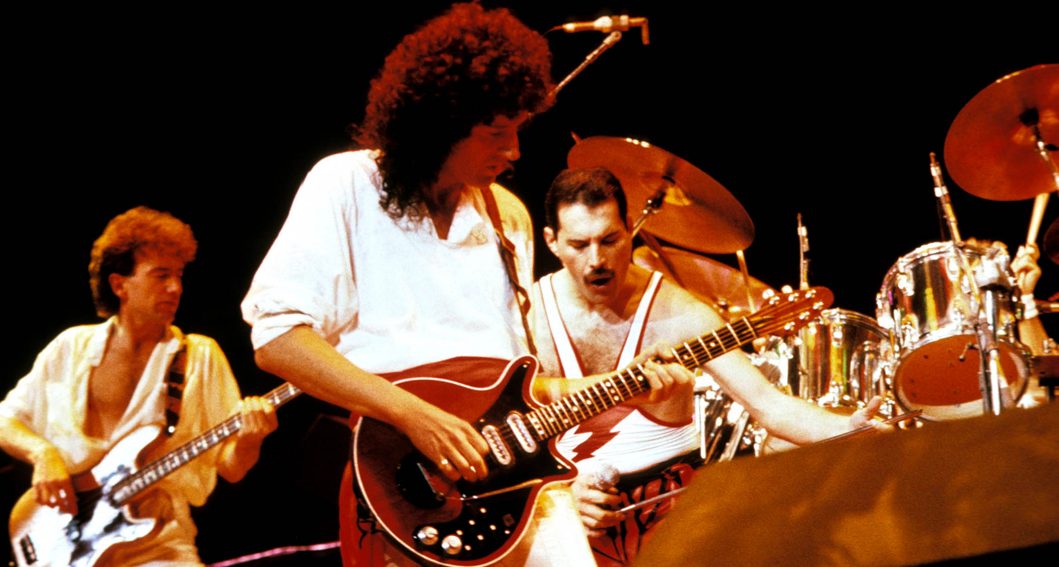 Queen perform in 1985 with guitarist Brian May in the foreground playing his iconic Red Special electric guitar. He wears all white.