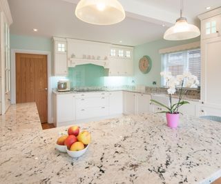 A general interior view of a white fitted shaker style kitchen with natural stone island, wicker bar stools, glass shade ceiling lights, potted orchid and fruit bowl