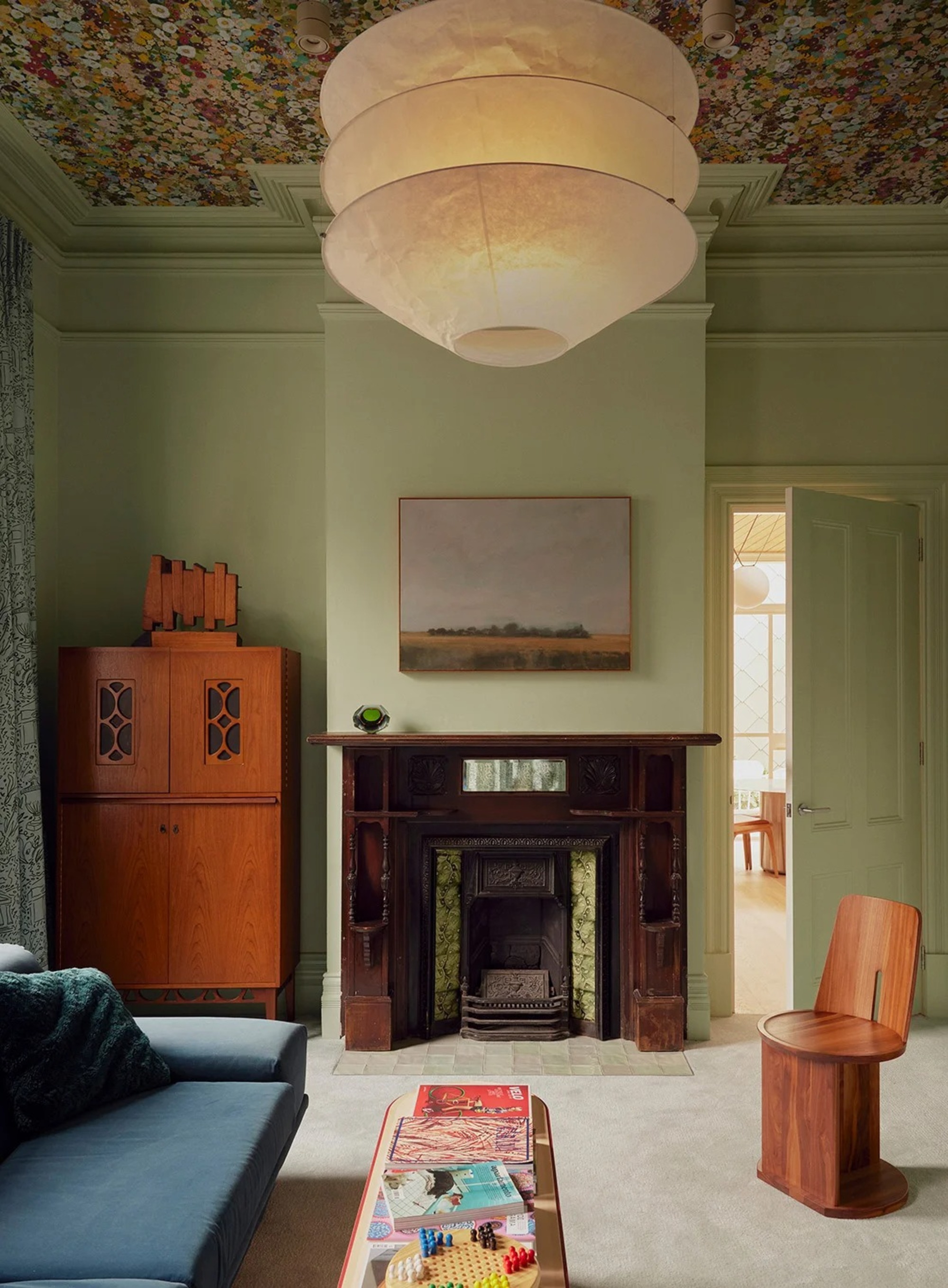 Image of a mint green living room with floral wallpaper on the ceilings, a lantern pendant light, a dark fire place, a blue sofa, and wooden furniture accents.