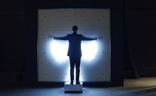 A man in a suit standing with both hands up on his side (shoulder lenght) on a metal square box in front of a large portrait frame that beams and mimics his posture to create an angel
