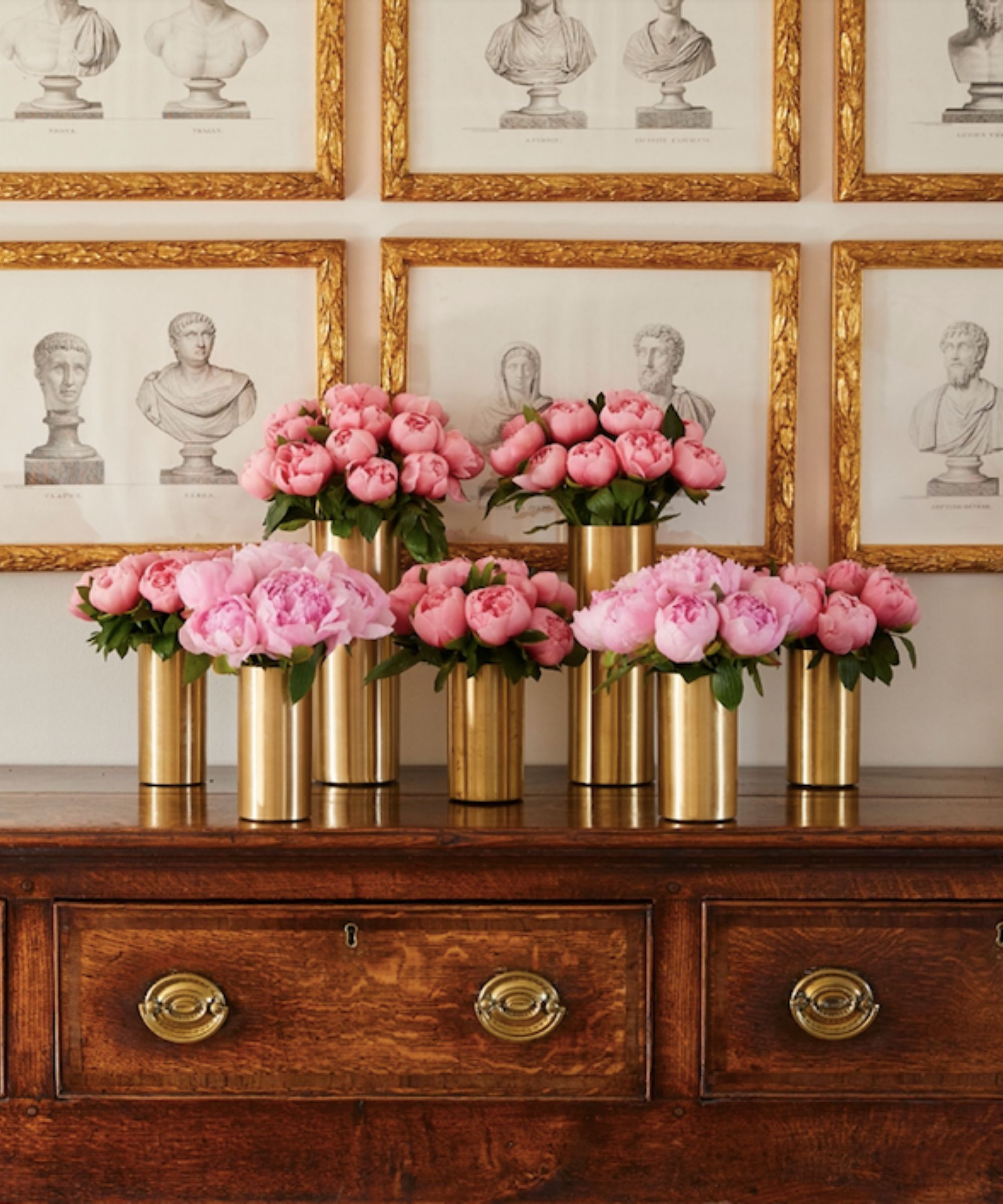 gold cylindrical vases filled with peonies at different heights on a dark wood sideboard with gold-framed prints behind