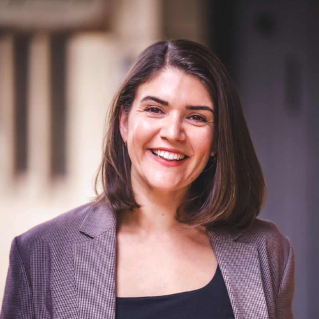 A woman with shoulder length brown hair wearing a blazer and smiling at the camera