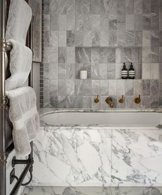 Mathroom with floor to ceiling gray marble tiles, smaller, dark gray marble tiles on wall, large light gray marble tiles on bath and floor, integrated alcove in wall for toiletries, metallic towel rail with white towels
