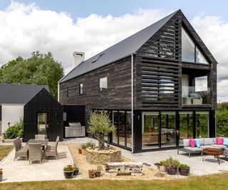 A large detached self build home clad in dark timber with a pitched roof and outdoor seating areas