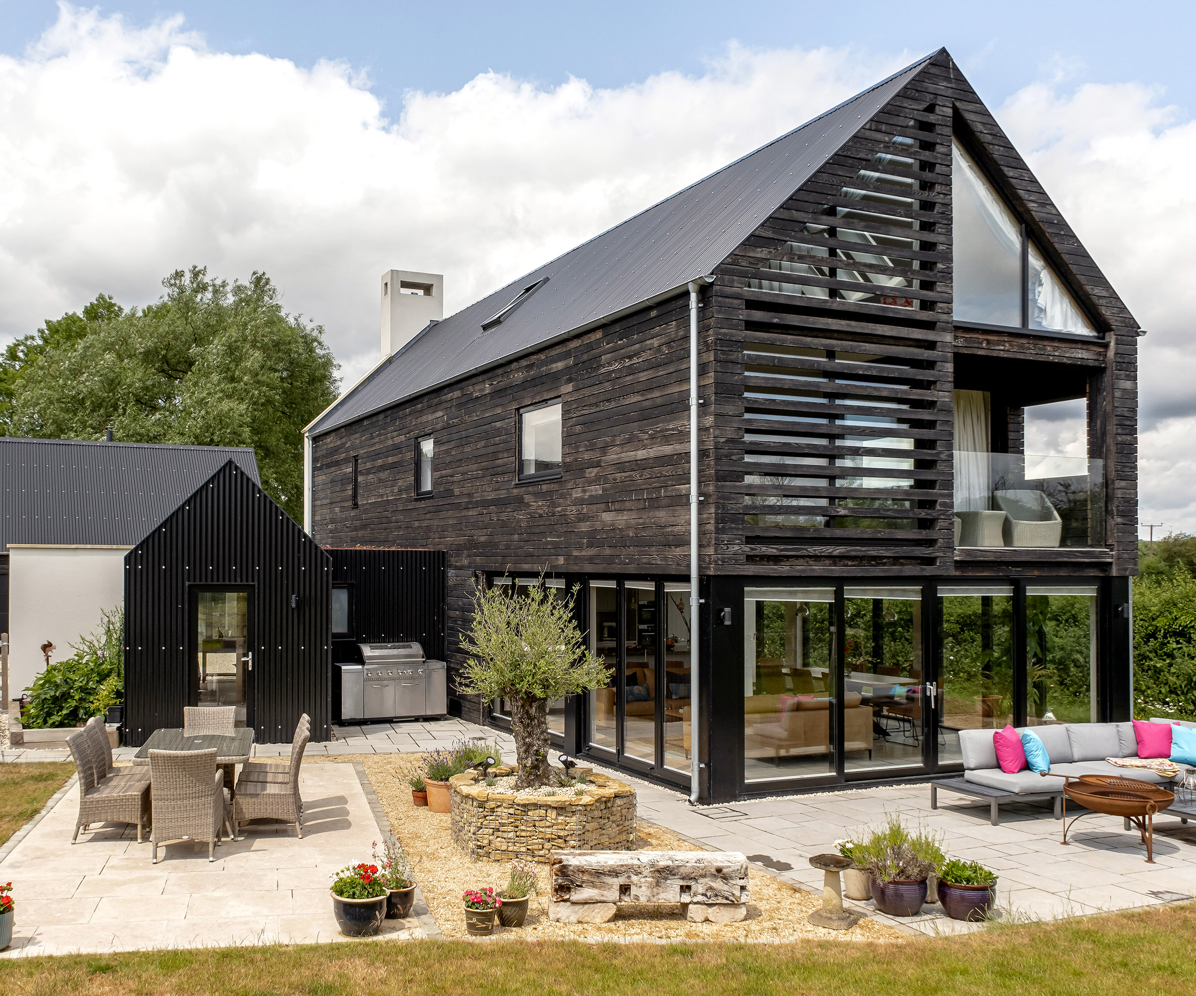 A large detached self build home clad in dark timber with a pitched roof and outdoor seating areas