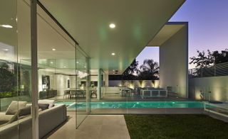 Exterior view of a residential white and glass home with a view of the pool, lawn, outside lounge area, inside lounge area and dining room.