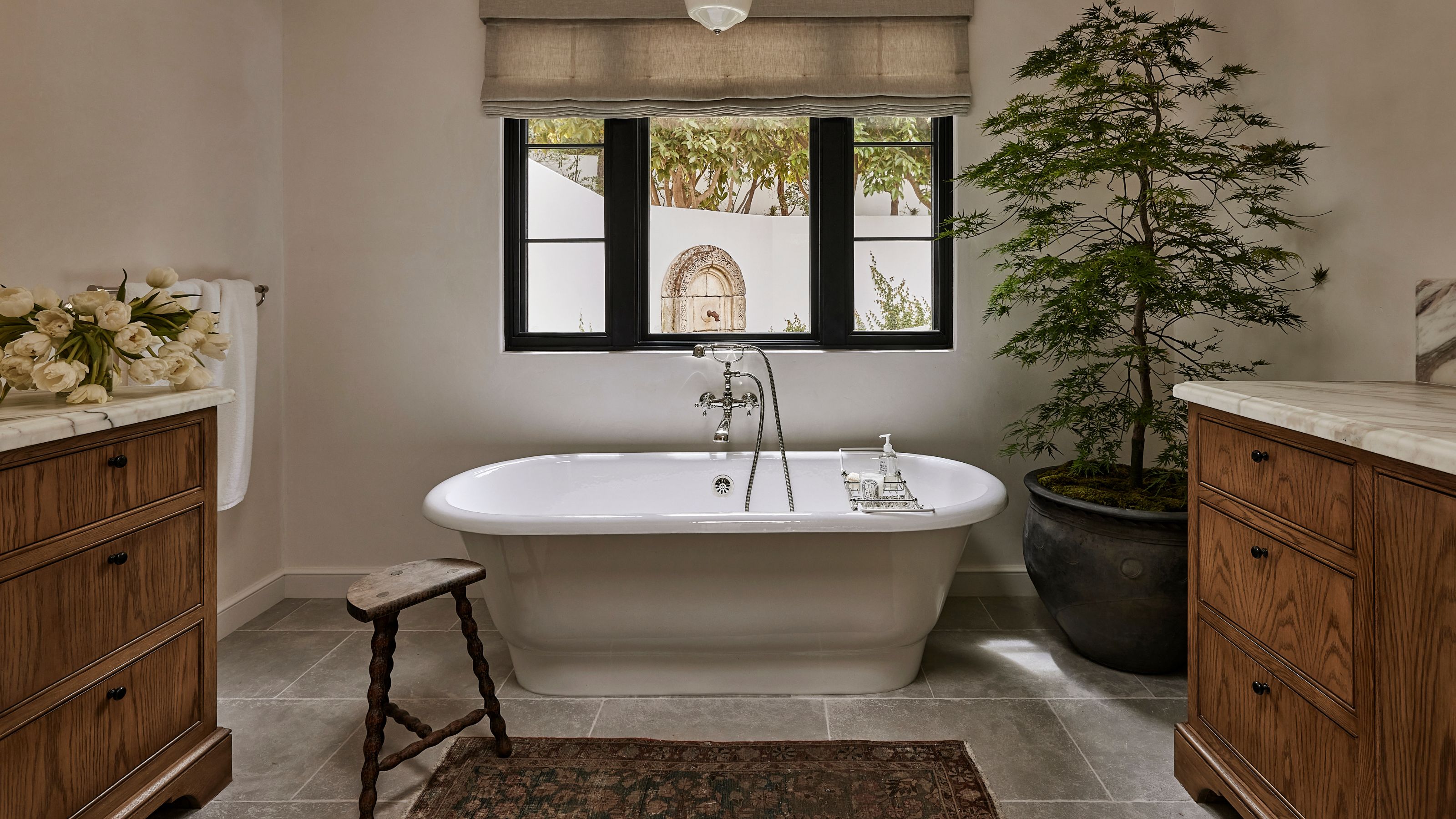 a large plastered neutral bathroom with warm wooden vanity units, stone floor and a freestanding tub beneath the window