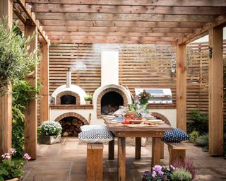Pergola ideas in a shaded area with wooden seating, outdoor kitchen and pizza oven and screened fence.