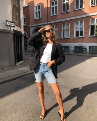 Model and jewelry designer Cecilie Moosgaard Nielsen poses on the streets of Copenhagen wearing black cat-eye sunglasses, a black blazer, white t-shirt, slim Bermuda denim cutoff shorts, chain bracelet, and strappy mule kitten heel sandals