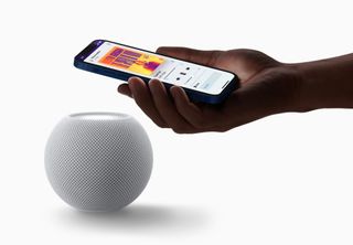En Apple Homepod mini smmen med en hånd som holder en mobiltelefon, sett mot en hvit bakgrunn.