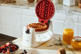‘Roto’ waffle maker. A white round waffle maker with a red inside cooking surface on a white marble kitchen counter.