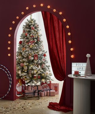 A hallway painted with red wall decor, with arched wall, set of string lights, hula hoops and pillar-effect side table with lightbox and Christmas tree