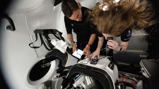 two people in black shirts float inside a cramped spacecraft capsule; one holds a small handheld device up to her eye