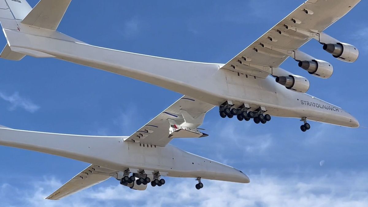 a large white twin-fuselage plane flies against a blue sky.