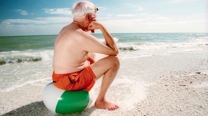 An older man sits on a beachball by the ocean dejectedly.