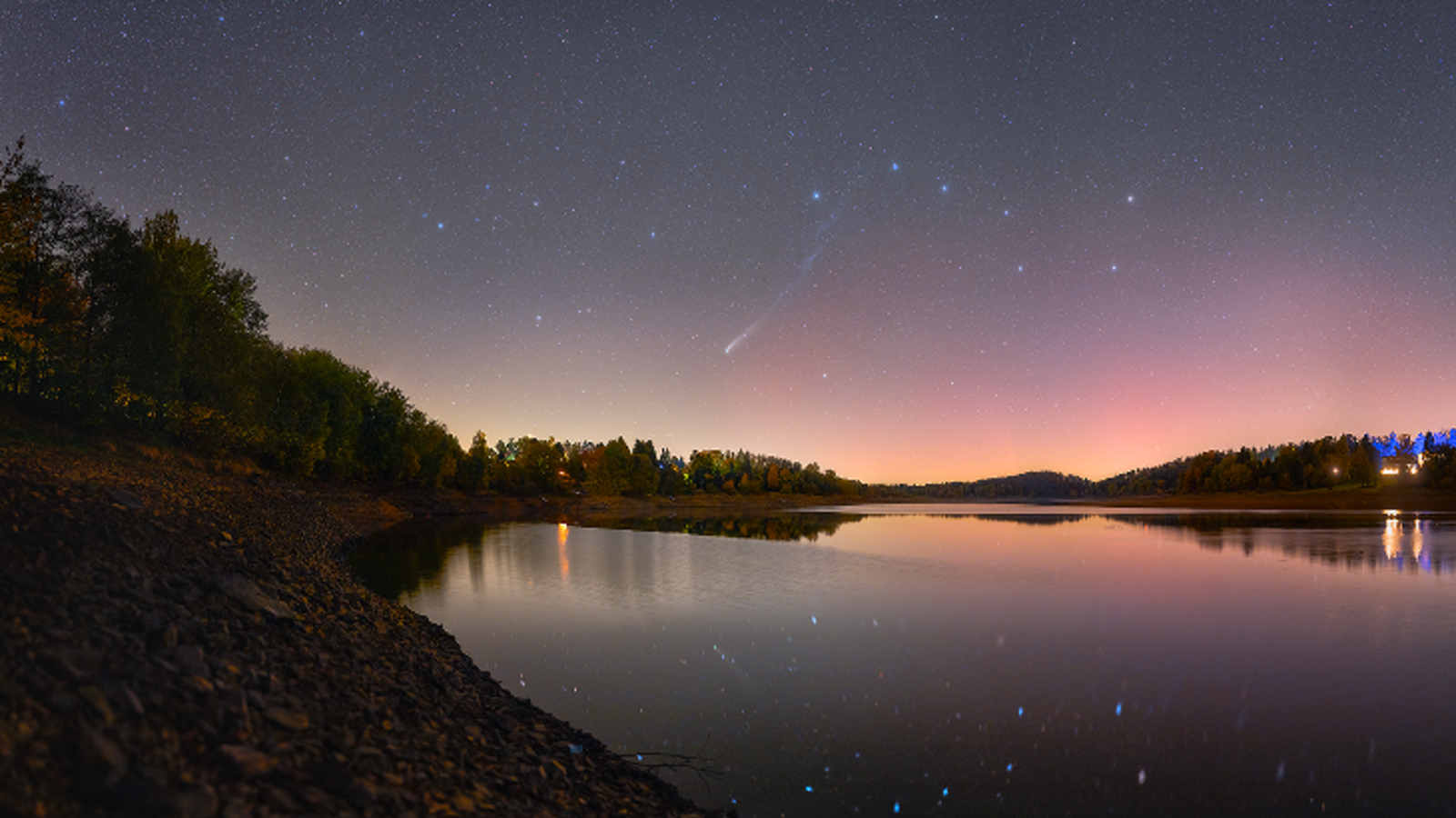 A photo of Comet Lemmon streaking across the night sky over a lake with faint red auroras