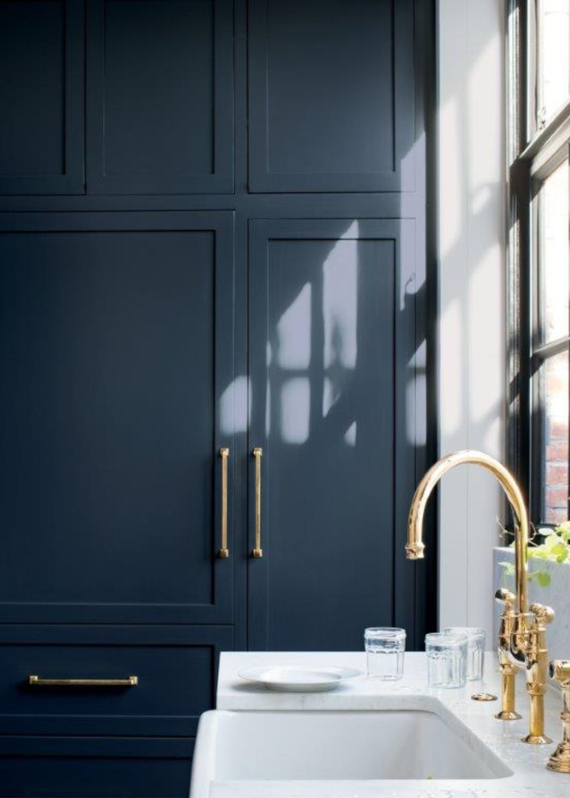 Corner of a modern kitchen with dark navy blue cabinets, white countertops with sink with brass fixtures, in front of a window