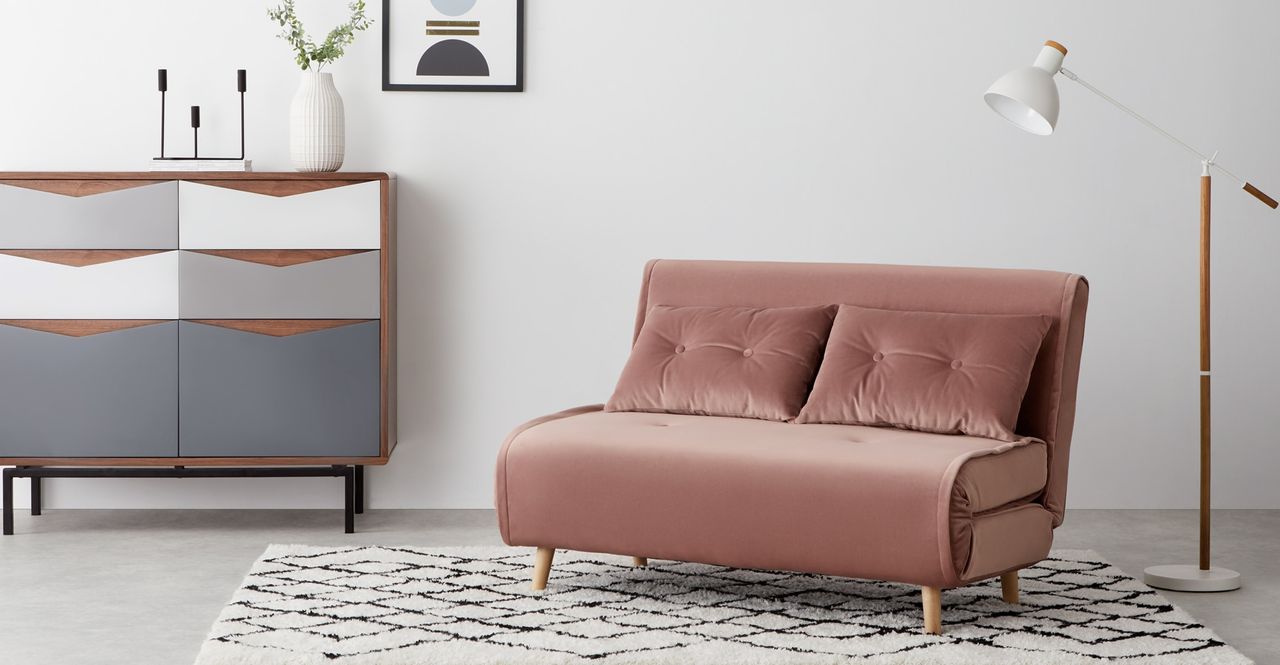 the Made Haru sofa bed in pink in a neutral living area, with white walls, a funky chest of drawers with white and blue drawers, a white and wood floor lamp, and some artwork on the wall