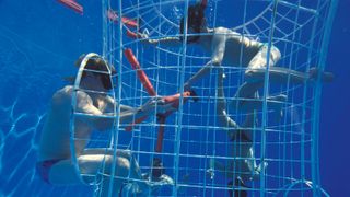 Two people swim in a metal cage in a pool