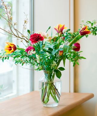 A vase of red and orange flowers with green stems in a clear vase, on a wooden ledge next to a window and beige walls