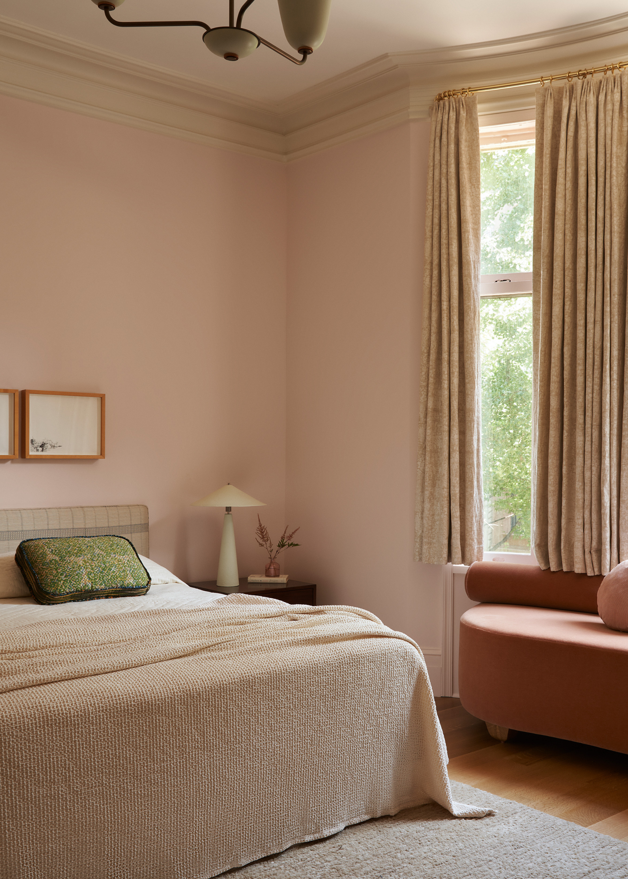 A beige bedroom with a tan couch, a natural bedscape, patterned pillows, a wood side table, a white table lamp, a vase of stems, and a pair of framed paintings