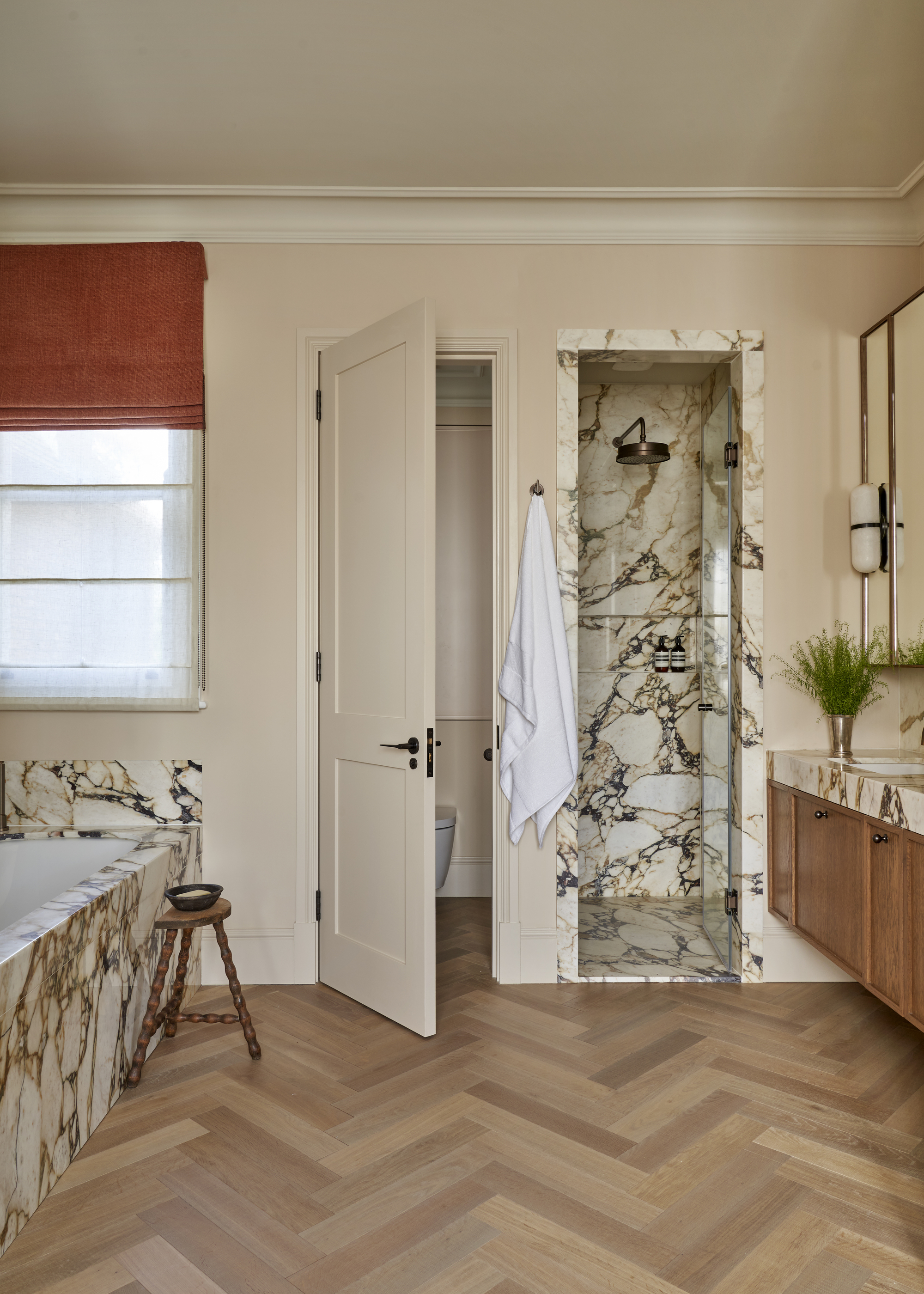 A bathroom with herringbone flooring, marble accents, and a walk-in shower with a marble door frame