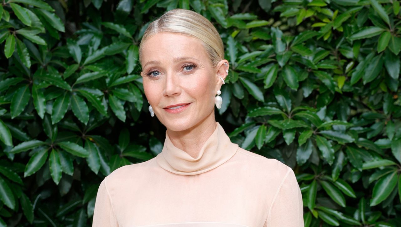 Gwyneth Paltrow standing in front of a wall of leaves wearing a blush tone sweater and large earrings