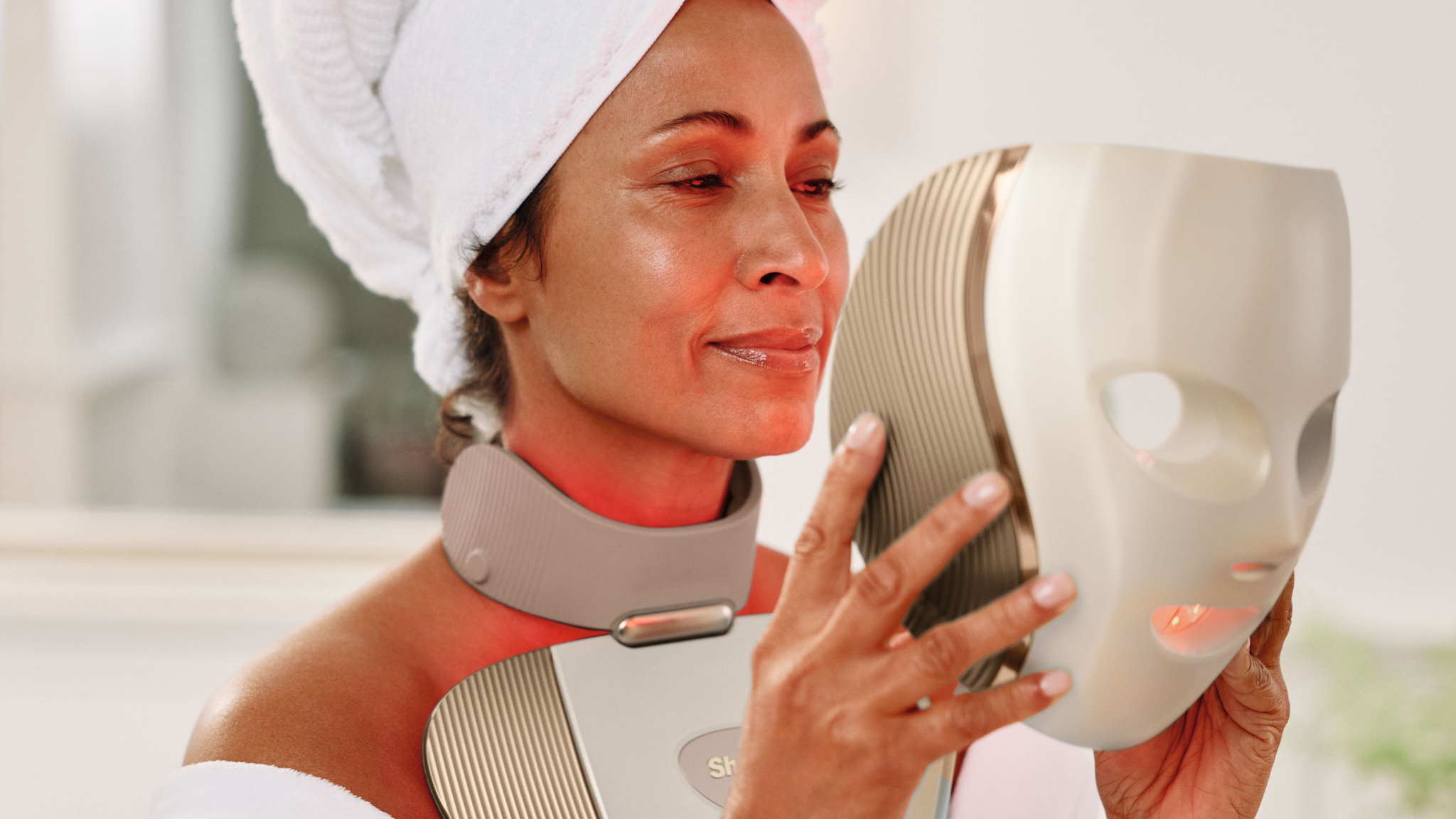 A woman with a white towel on her head puts on the Shark Beauty CryoGlow Clinic Face + Body mask