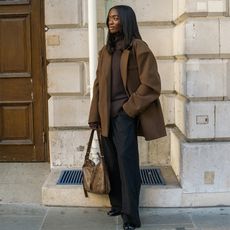 liv wears brown sweater, brown coat, black pants, black tabbis, and brown handbag while posing on the street