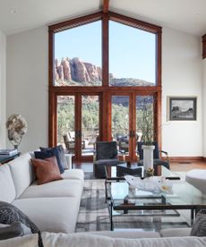Large living room with floor to ceiling windows with wooden frame, looking out to a mountain view. Cream sofa, gray armchairs, glass coffee table in foreground