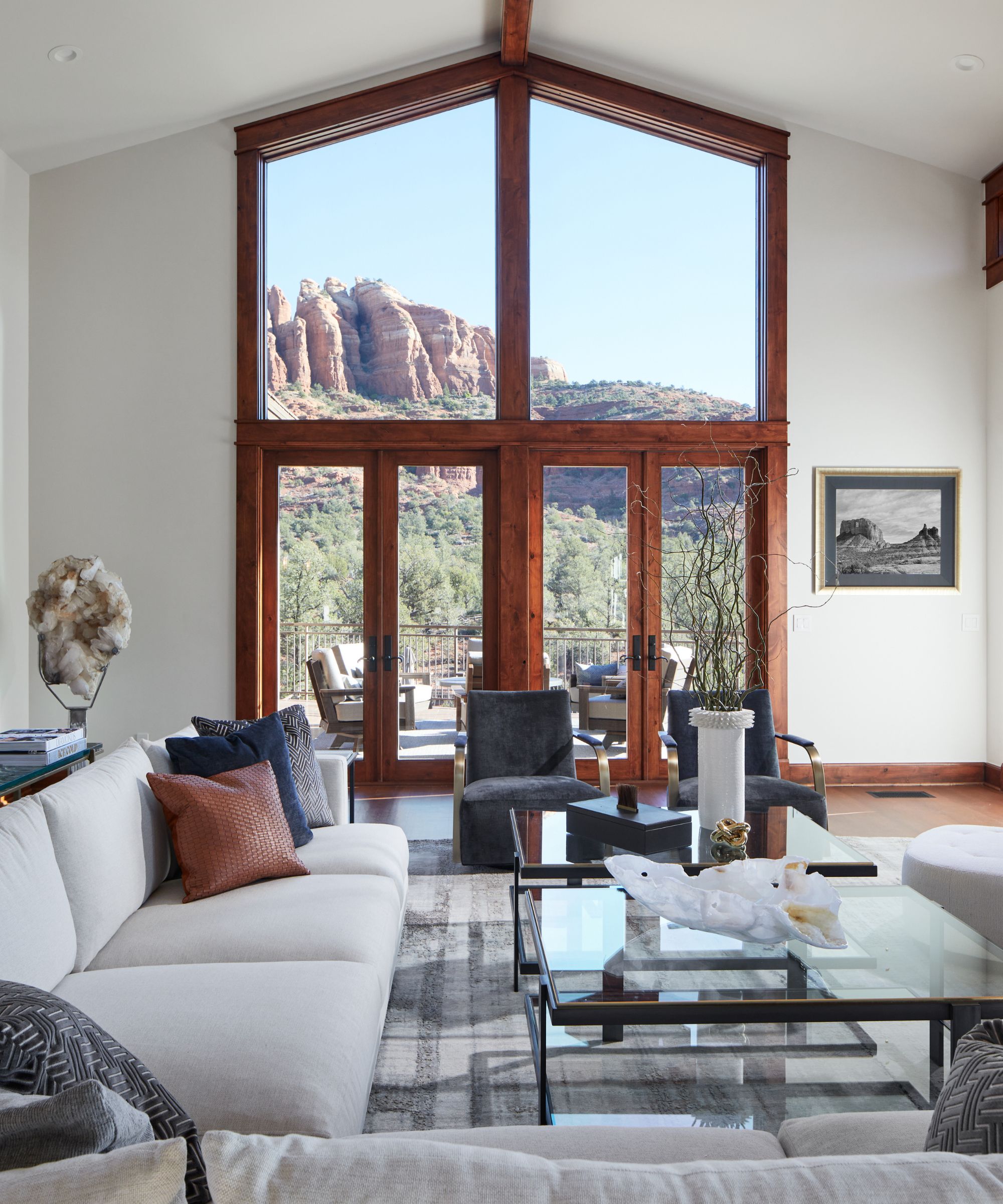 Large living room with floor to ceiling windows with wooden frame, looking out to a mountain view. Cream sofa, gray armchairs, glass coffee table in foreground
