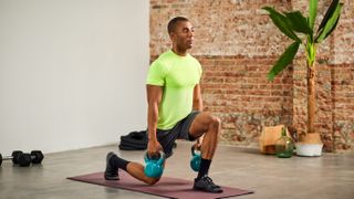 A man in sportswear performs weighted lunges. He holds a pair of kettlebells in his hands and both knees form right angles; the front one is elevated and the back one grazes the floor. In the background, we see a red brick wall, a plant and some dumbbells.