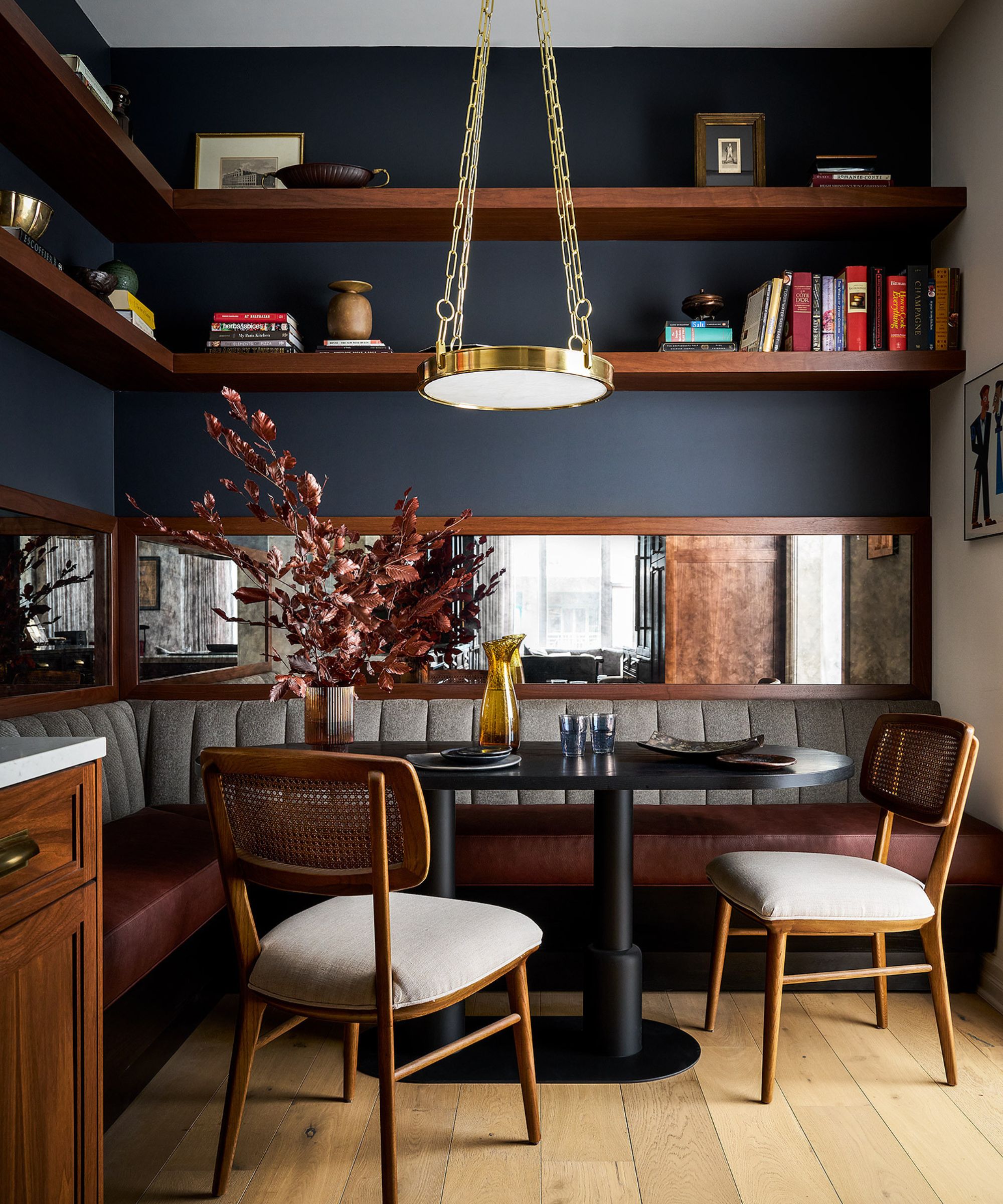 A cozy dining nook with dark blue walls, dark wood open shelving, and a mirrored wall above the banquette seating, with a wooden edge. A black modern dining table, two wooden chairs, and hardwood flooring.
