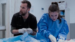 Noah Wyle holding a medical instrument and looking to his left while Isa Briones does a medical procedure. 