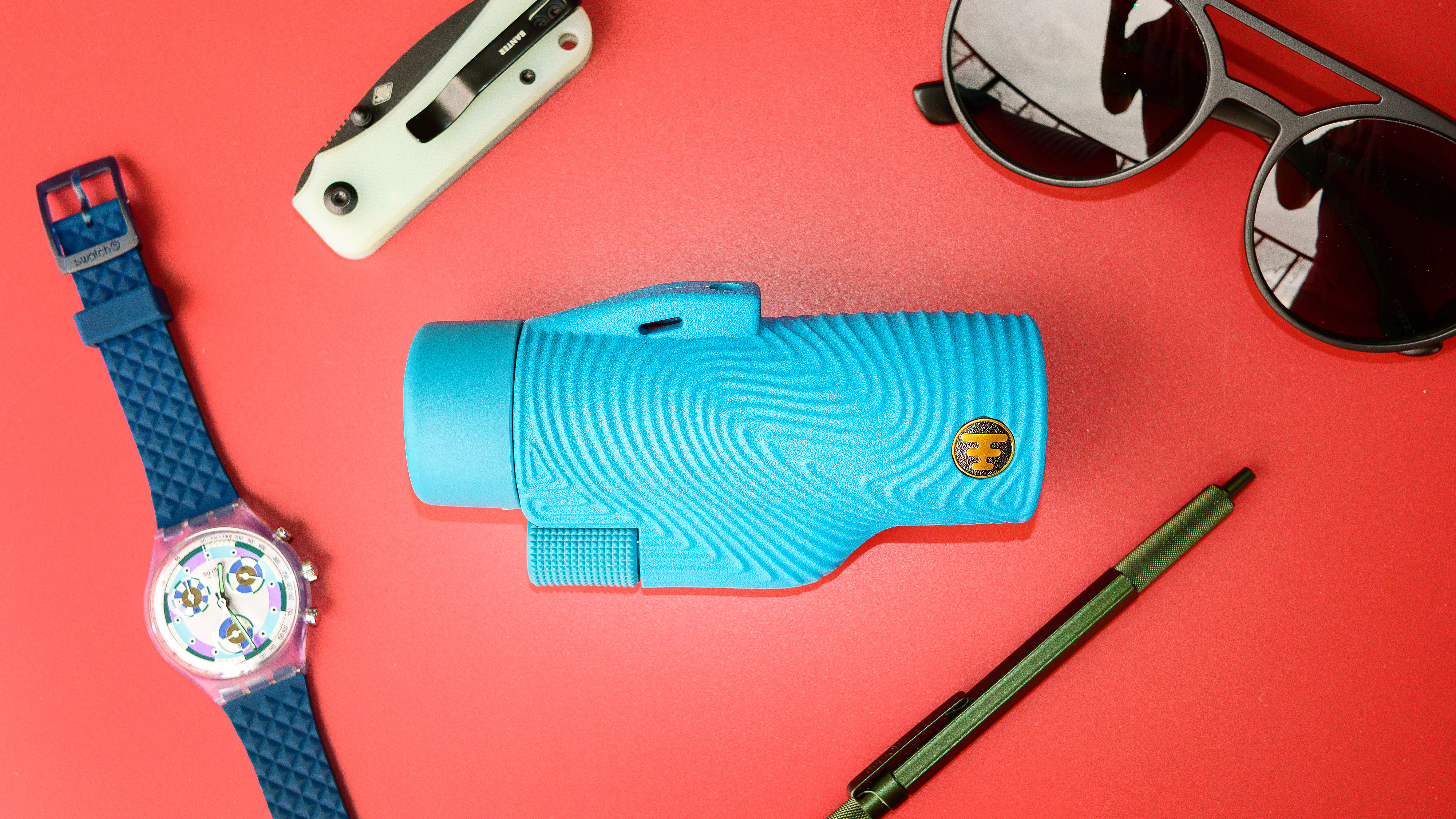 A blue Nocs Field Tube monocular on a red table surrounded by a Garmin watch, a pocket knife, sunglasses and a pen.