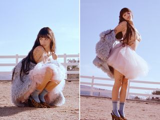 Singer Rebecca Black on a farm. She is standing in a dirt lot in front of white fence wearing a pink lace bra styled with a pink tutu skirt, shaggy gray coat, gray socks, and leopard-print slingback heels.