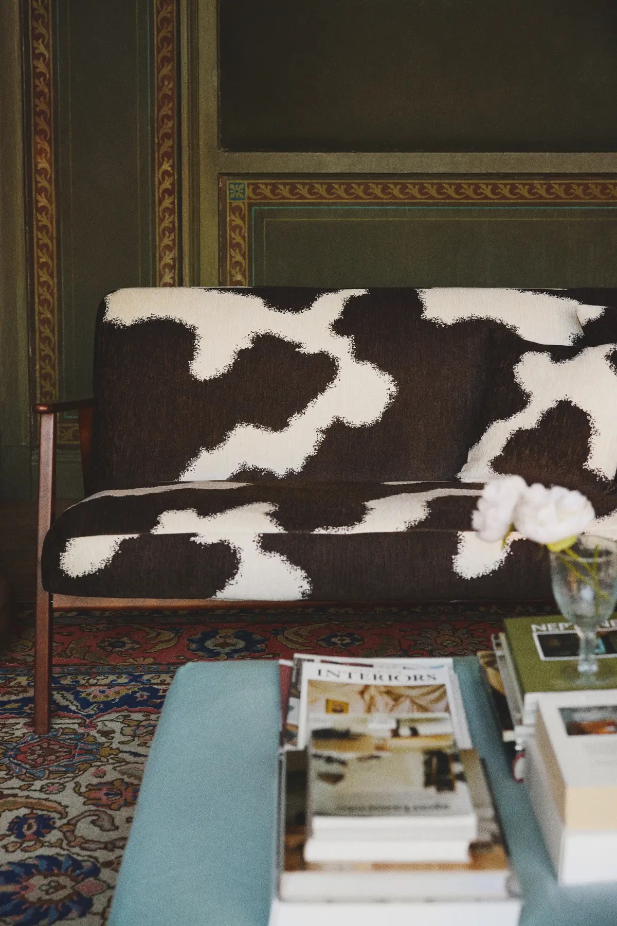 Image of a brown and white, cow print-looking sofa with a traditional red and blue rug underneath and a blue coffee table with stacks on magazines on it.